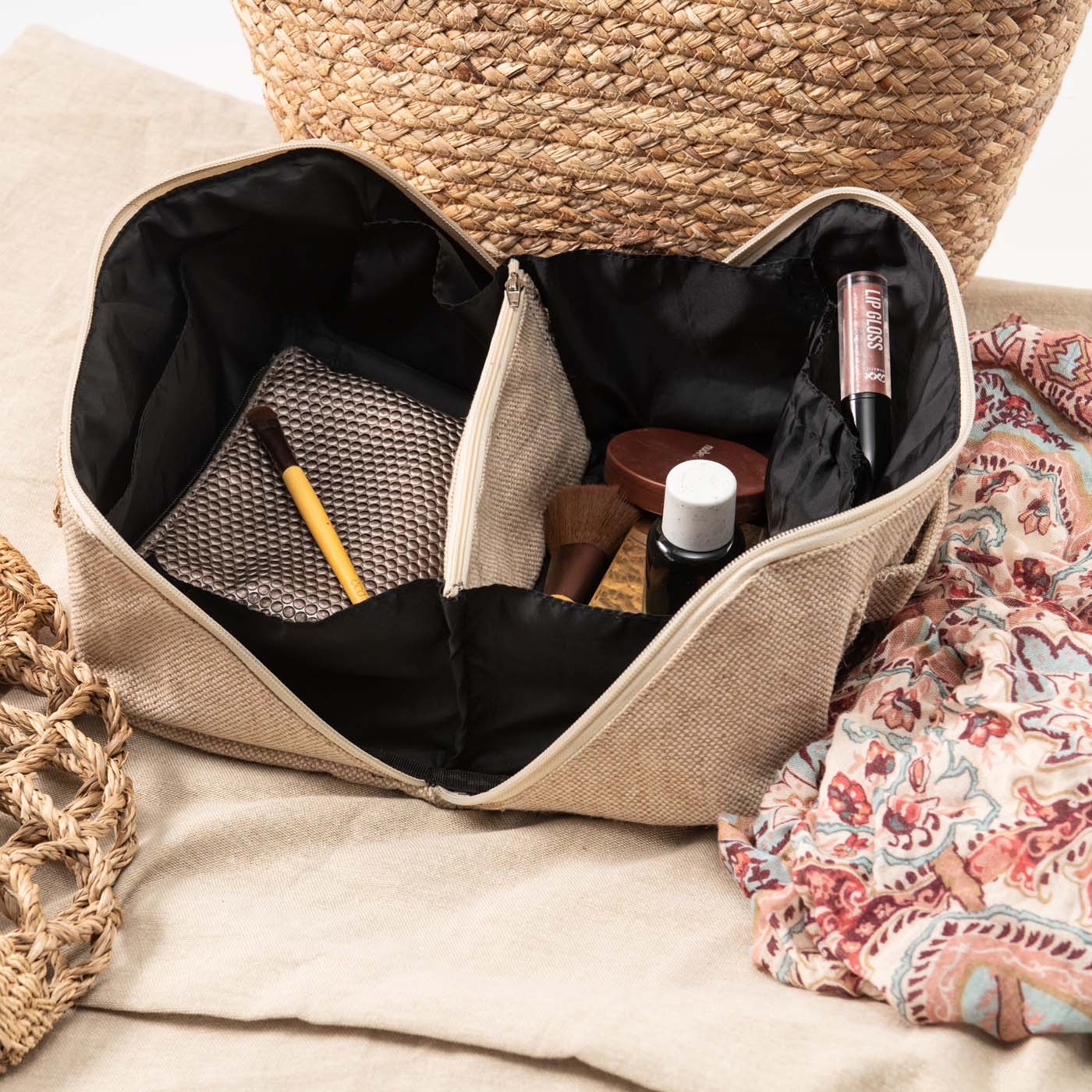 A beige flat lay travel makeup bag with an open flat design, featuring a divider and a handle, displayed on a flat surface with a patterned fabric in the foreground.
