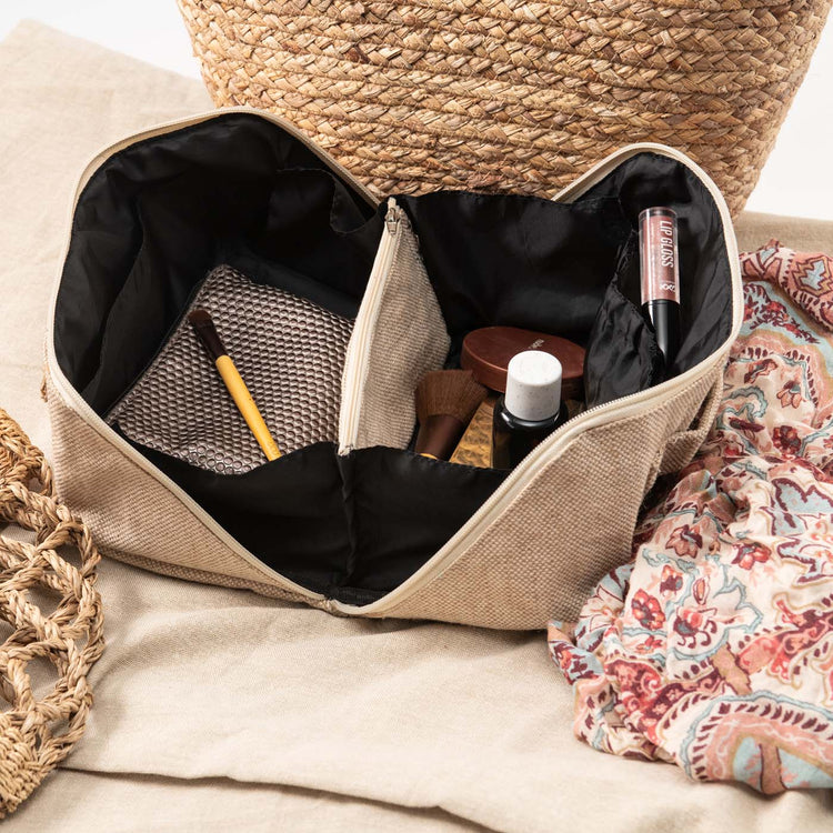 A beige flat lay travel makeup bag with an open flat design, featuring a divider and a handle, displayed on a flat surface with a patterned fabric in the foreground.