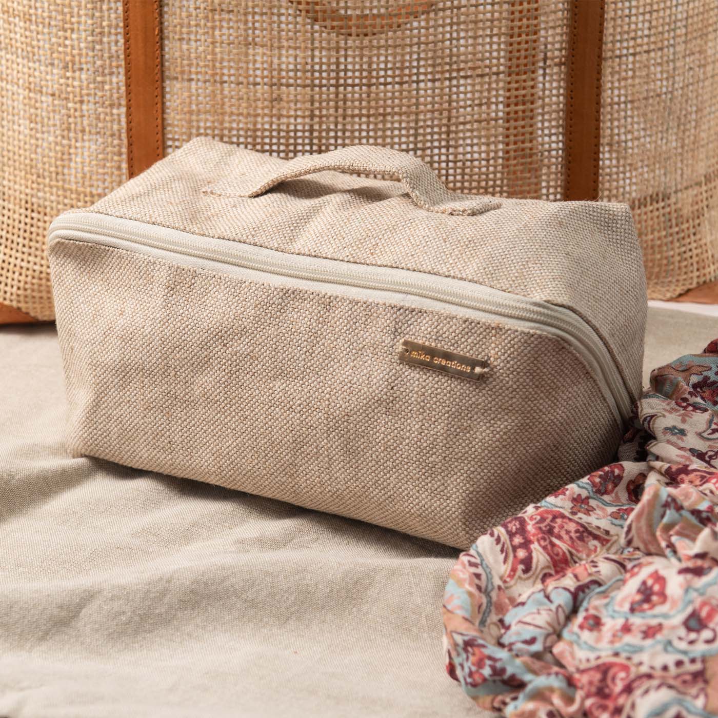 A beige travel makeup bag with an open flat design, featuring a divider and a handle, displayed on a flat surface with a patterned fabric in the foreground.